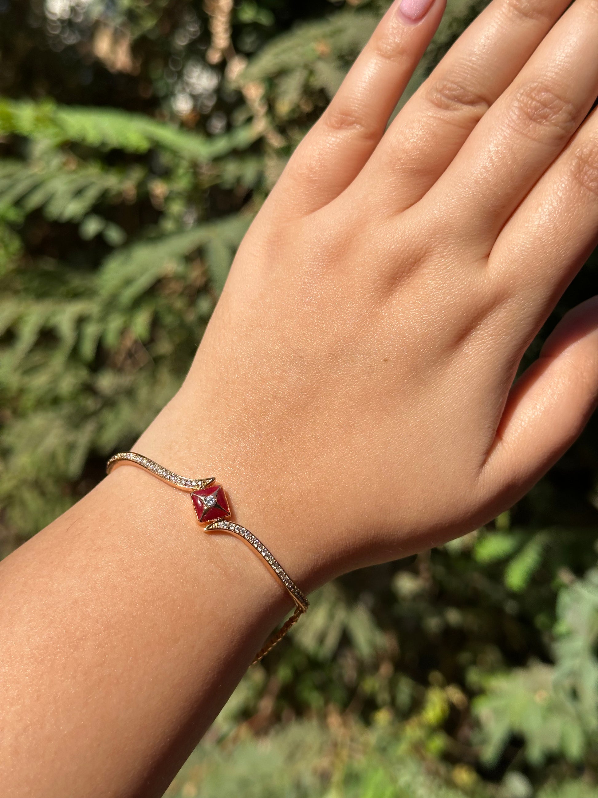 A gold bracelet with a diamond clasp on a person's wrist, displayed against a leafy background.